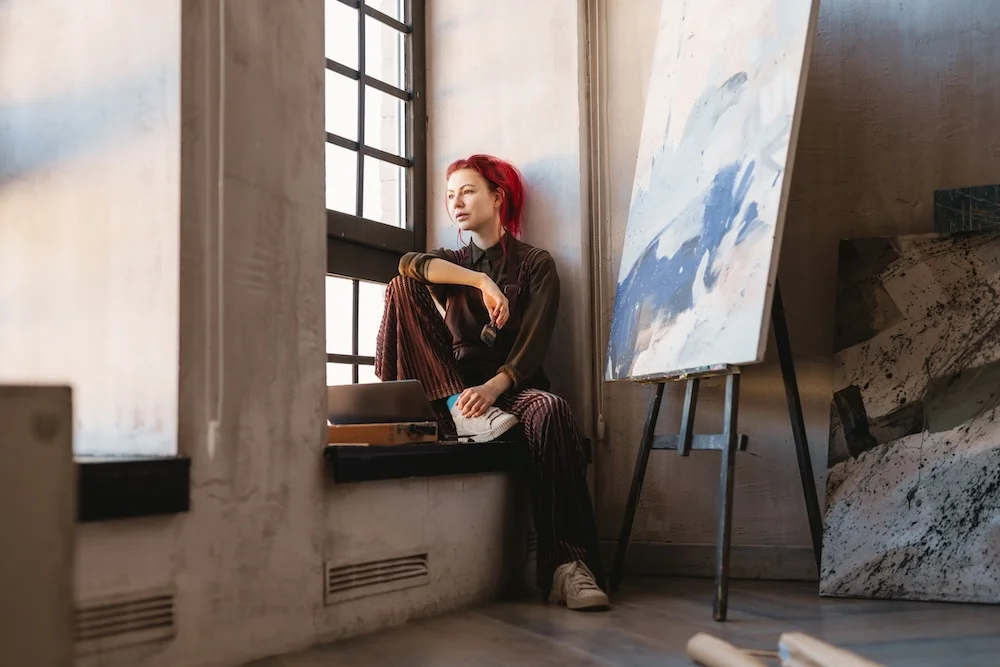 Person sitting on a wide windowsill in an art studio with large windows, next to an easel holding an abstract painting and additional canvases leaning against the wall.