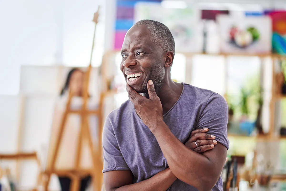 Person standing indoors with arms crossed, wearing a casual t-shirt, in a bright art studio with easels and colourful paintings in the background.