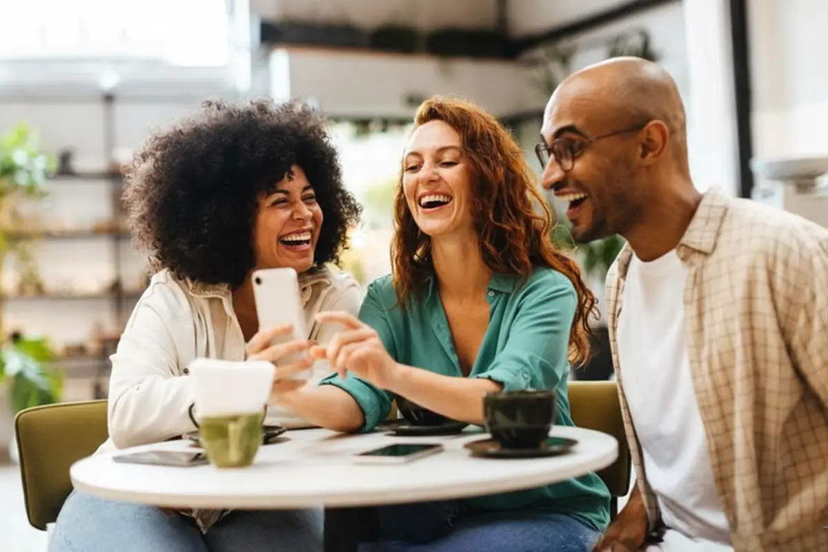 Friends laughing together while using a mobile phone at a café.