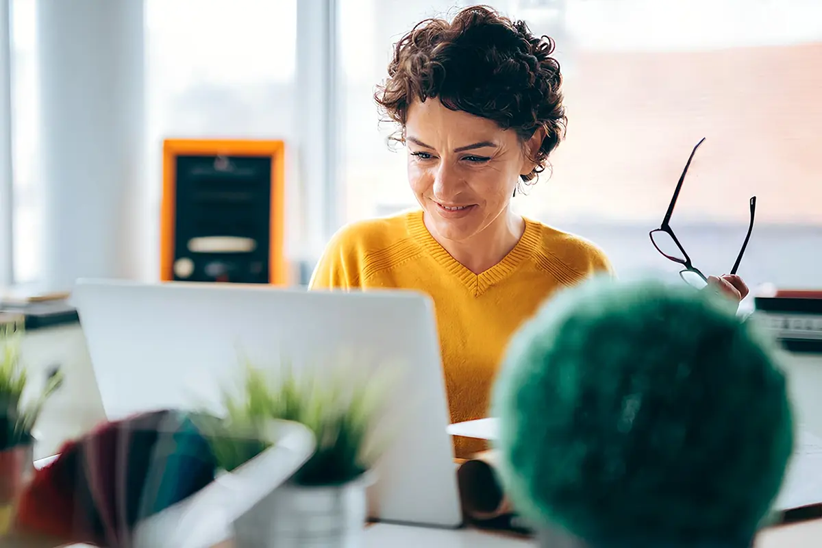 Working in a vibrant, creative office space. Person sitting at a desk with a laptop, holding eyeglasses in one hand, surrounded by green plants and colourful design materials in a bright workspace.