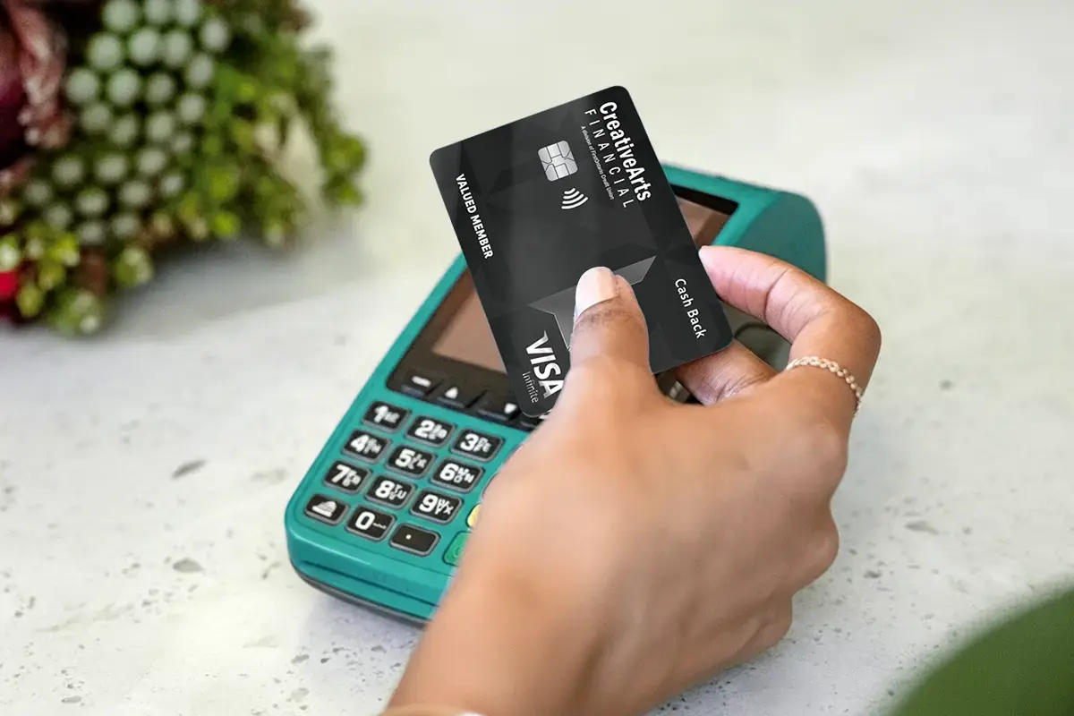 Hand holding a Creative Arts Financial Visa credit card near a teal card reader for a contactless payment.