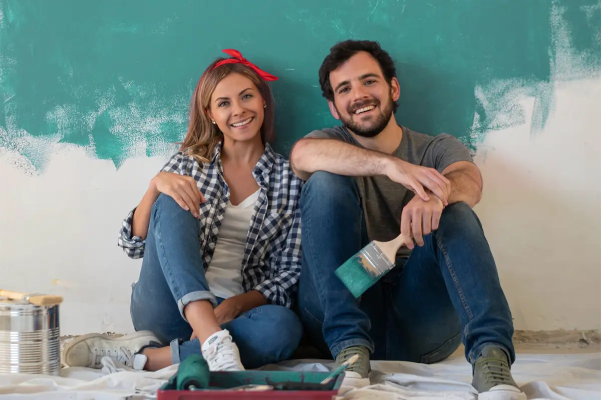 A happy couple sit on the floor in front of a partially painted teal wall.