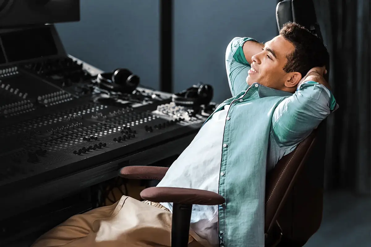 Relaxing in a modern music production studio. Person leaning back in a chair with hands behind the head in a professional audio recording studio, surrounded by a large mixing console and headphones.