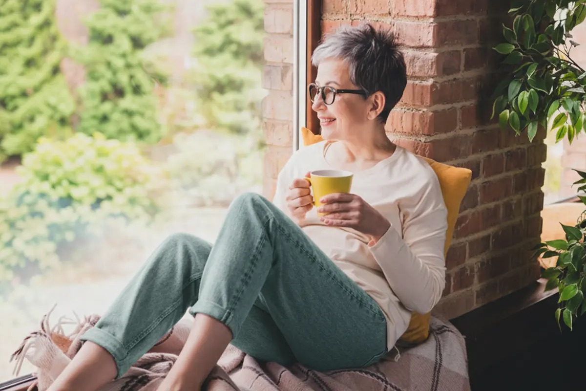 Woman sitting by a window holding a yellow coffee mug.
