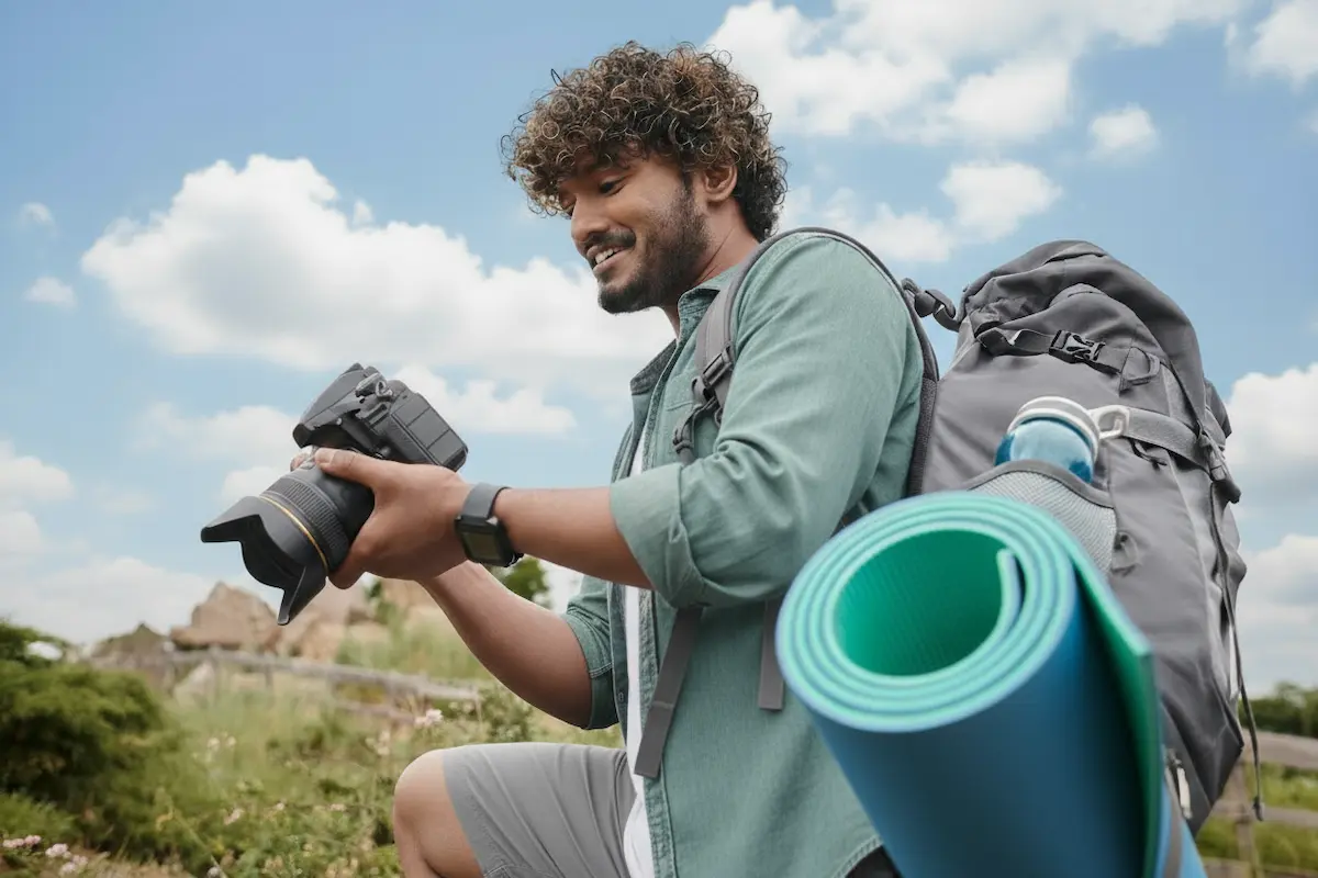 Person outdoors holding a DSLR camera with a large lens, wearing a green shirt and carrying a gray backpack with a rolled-up blue yoga mat attached.