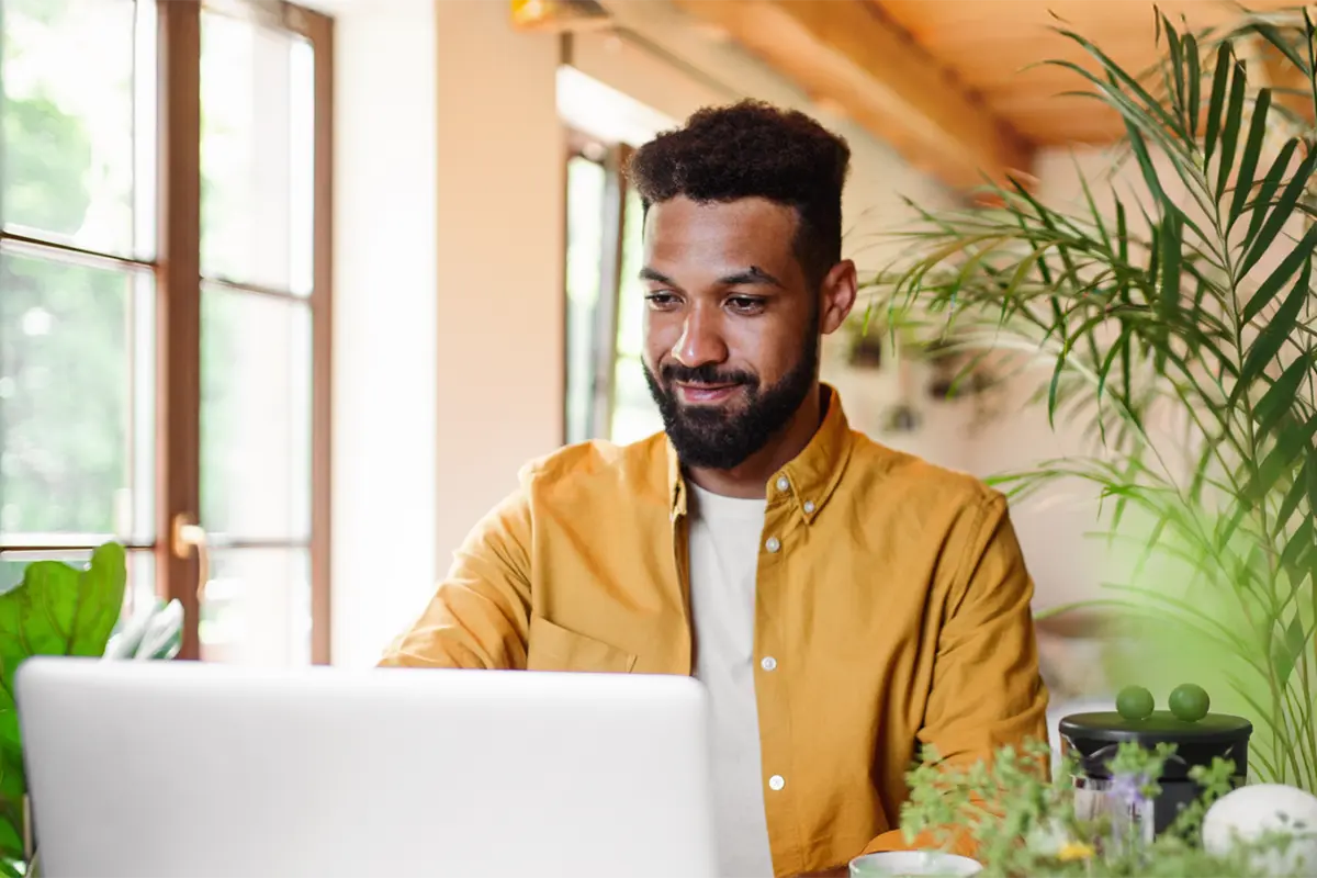 Man working on a laptop at a bright desk surrounded by plants.