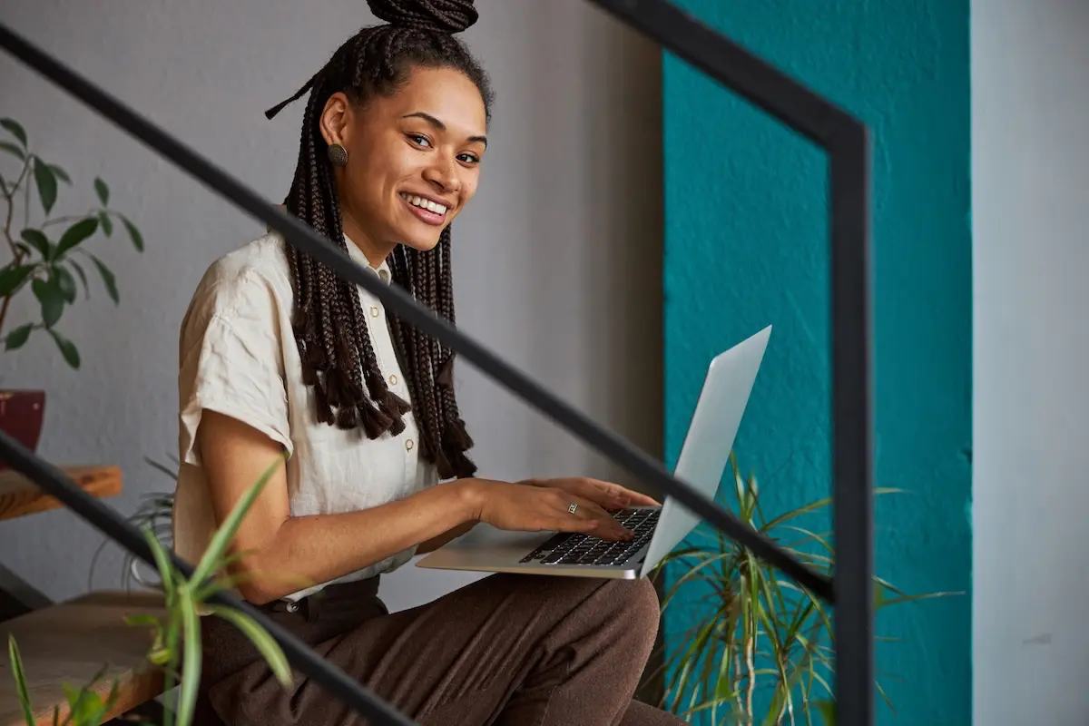 A smiling Black woman with braids works on her laptop while sitting on stairs in a modern space, illustrating a remote freelancer confidently building financial security through smart budgeting.
