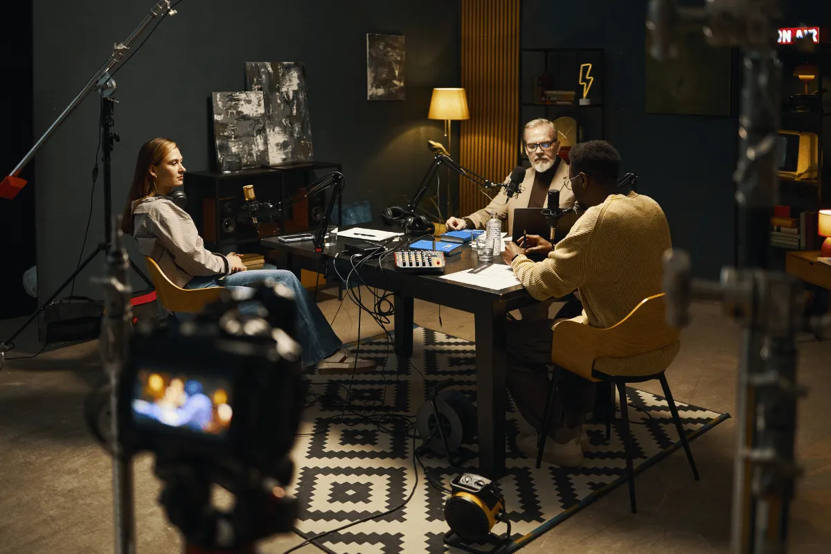 Three individuals recording a podcast around a table with microphones, headphones, and audio equipment in a studio.