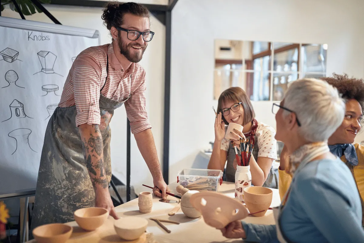 Instructor leading a pottery class, showing clay work to a group seated at a table with bowls, tools, and sketches visible.