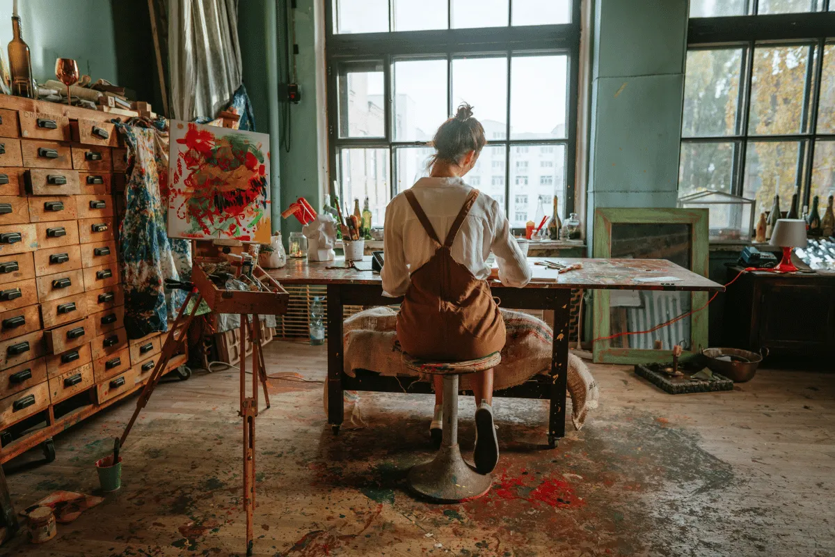 Artist working at a desk in a paint-filled studio, surrounded by canvases, brushes, and scattered materials near a large window.