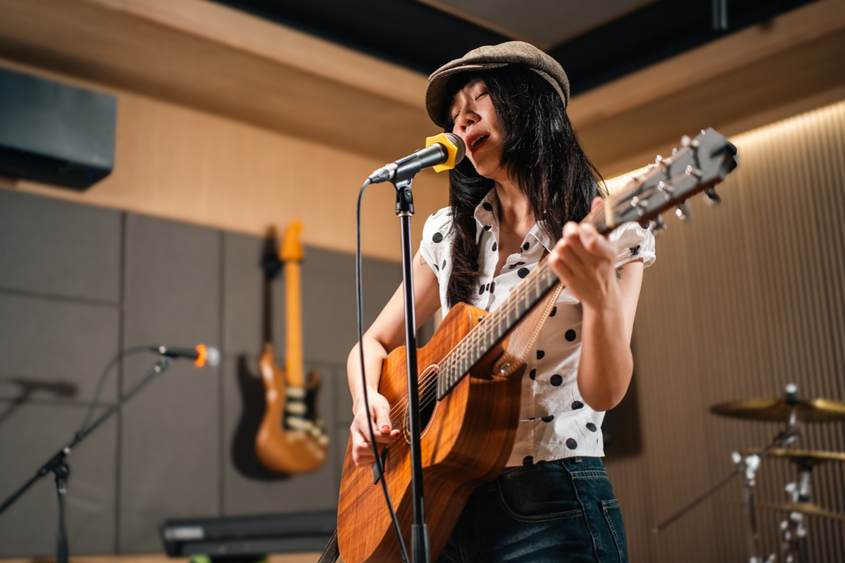Musician singing into a microphone while playing an acoustic guitar in a recording studio.